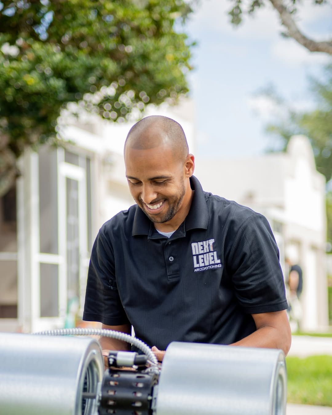 Smiling technician in a black branded polo assembling an inline duct fan between two duct sections, outdoors in daylight